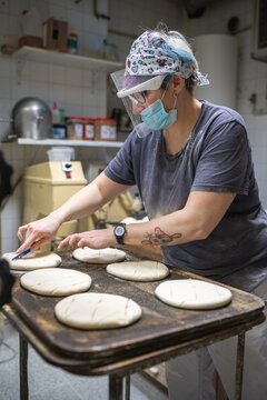 Female baker wearing mask and shield standing at table and making notches on raw dough before baking