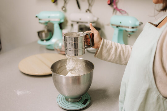 Cropped unrecognizable professional female confectioner sieving flour into metal bowl while preparing dough for sweet pastry in kitchen