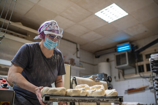 From below of female baker in mask and shield standing at table in kitchen and making round shaped bread from raw dough while working in bakehouse