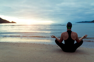 Back view of unrecognizable swimmer in swimsuit and cap sitting in Lotus pose with mudra hands on seashore and doing yoga at sunset