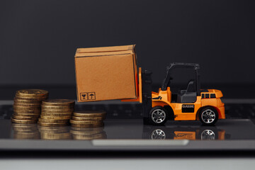 Orange toy forklift with carton boxes and coins on keyboard close-up. Logistics and delivery concept.