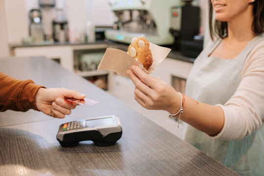 Side view of female seller holding delicious sweet cookie while customer paying with credit card using contactless machine in confectionery cafe