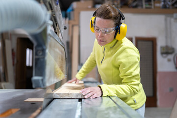 Side view of focused adult female artisan in goggles and headphones sawing wooden board with sharp circular saw in workshop