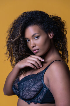 Attractive African American overweight female model with long curly hair wearing delicate lace bra looking at camera against yellow background