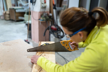 Side view of anonymous female carpenter in uniform and goggles using sharp saw while cutting wooden plank in workshop