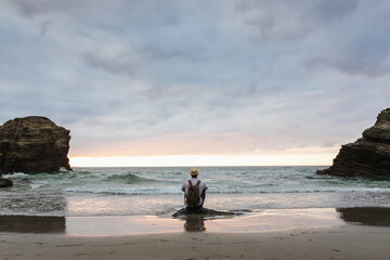 Back view of anonymous male traveler sitting on a rock at sandy coast enjoying views during summer vacation near sea at sunset