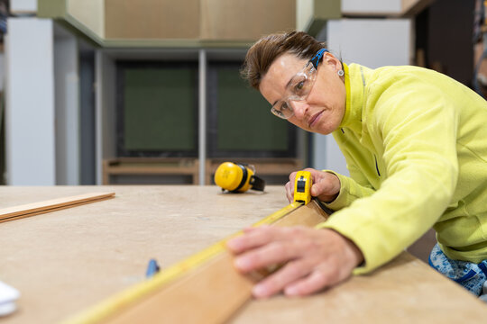 Low angle of concentrated middle aged female carpenter in uniform and protective goggles measuring wooden plank with roulette during work at table