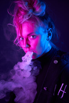 Side view of young cool female smoking vape in neon light in dark studio looking at camera