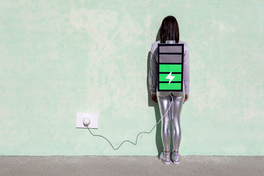 Back view of unrecognizable female with long dark hair in stylish outfit standing on street near wall with battery shaped backpack connected to charger