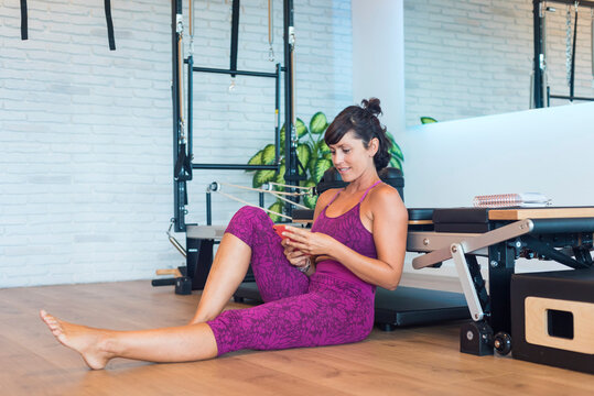 Happy female instructor sitting near Pilates reformer and dialing phone number from notebook on smartphone in gym
