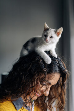 Side view of smiling female owner with white funny adorable little kitten on head resting at home