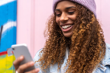 Cheerful African American female teenager in trendy outfit and hat smiling while reading message standing on street near pink door