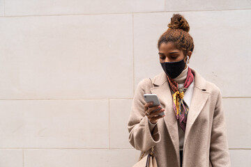 Trendy young ethnic woman in elegant coat and face mask with bag standing near wall and talking on smartphone using wireless earphones