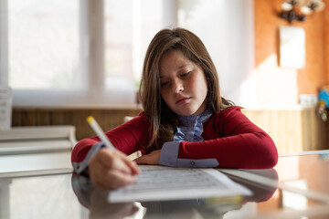 Concentrated schoolgirl in casual clothes siting at table and writing on paper during exam preparation at home in sunlight