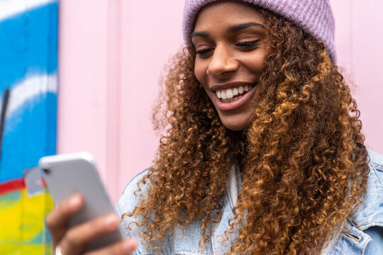 Cheerful African American female teenager in trendy outfit and hat smiling while reading message standing on street near pink door
