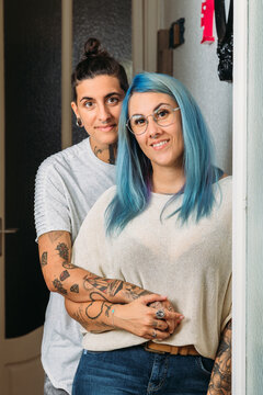 Androgynous tattooed woman embracing girlfriend with dyed hair from back and looking at camera while resting in hallway at home