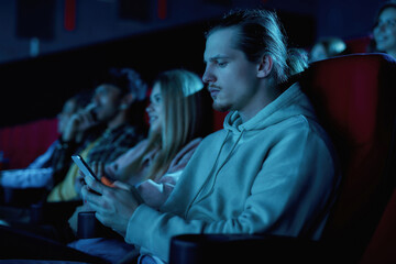 Fototapeta premium Focused young guy using his smartphone while missing boring movie at the cinema