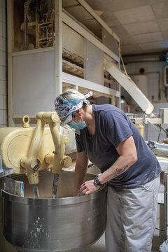 Side view of female baker wearing mask and shield taking piece of raw dough from kneader while working in bakery