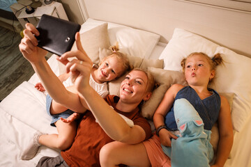 Mom and her two daughters are taking selfie on the smartphone relaxing on bed.