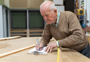 Concentrated elderly male artisan in casual clothes taking notes while working in joinery