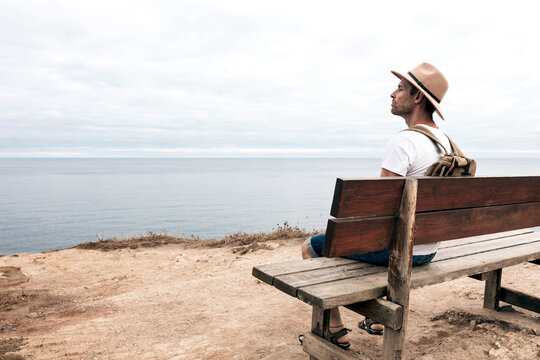 Side view high angle of male explorer in hat sitting on wooden bench and admiring spectacular scenery of sea and rocks