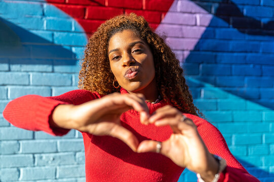 Confident young ethnic lady with Afro hair in casual wear pouting lips and showing heart sign with hands while standing on street near brick wall with colorful graffiti