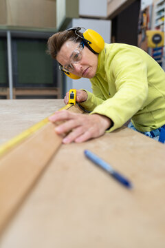 Low angle of concentrated middle aged female carpenter in uniform and protective goggles measuring wooden plank with roulette during work at table