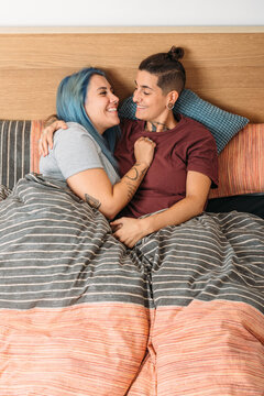 From above tattooed androgynous woman cuddling happy girlfriend with dyed hair while resting on bed at home together looking at each other