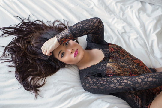 From above of slim sexy female wearing black lace bodysuit lying on bed and looking at camera