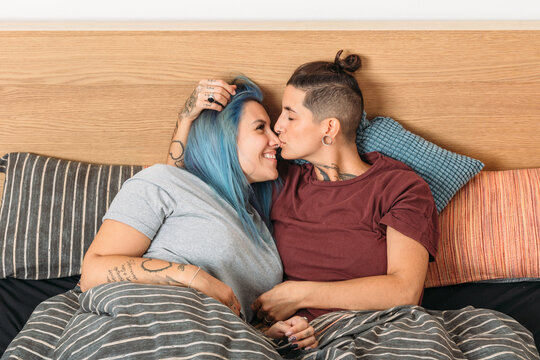 Tattooed androgynous woman kissing happy girlfriend with dyed hair while resting on bed at home together
