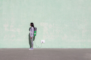 Side view of young fit female in trendy outfit standing on street with creative backpack connected to charger and having phone conversation
