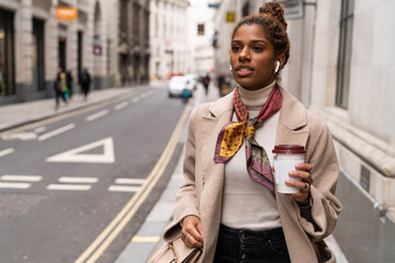 Confident young African American female entrepreneur in fashionable outfit listening to music with wireless earphones and drinking takeaway coffee while walking on city street during break