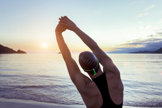Back view of fit male swimmer in swimsuit and cap standing on seashore and stretching arms before training - Powered by Adobe