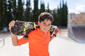 Adorable pensive child in casual pink t shirt looking at camera while standing in skate park and holding board behind head
