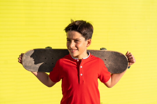 Happy teenage boy holding skateboard while standing in yellow background looking away