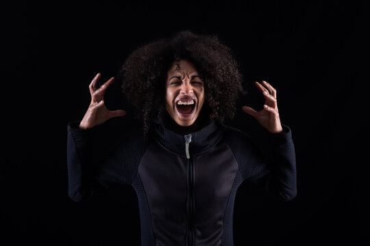 Stressful angry young African American female with curly hair shouting loudly and touching head while standing against black background