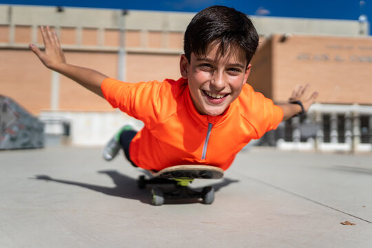 Cheerful ethnic teen boy in casual clothes lying on skateboard with outstretched arms and smiling during training on square on sunny day