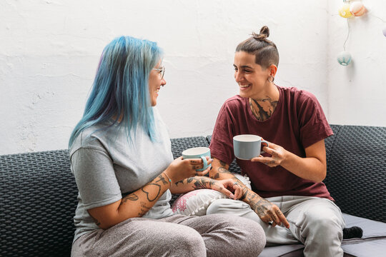 Delighted lesbian couple with mugs of hot beverages smiling and looking at each other while relaxing on couch at home together