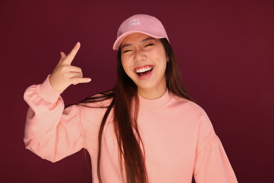 Portrait of happy young asian woman in the studio wearing pink clothes over garnet background. She is winking and making the rock sign