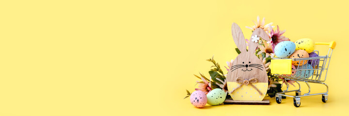 Easter composition with colorful eggs in shopping cart, wooden bunny and spring flowers on yellow background. Banner. Copy space.