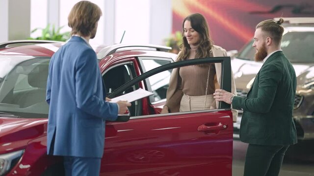 Young Elegant Caucasian Man With Red Beard Opening Car Door For Beautiful Woman As Car Dealer Talking At Front. Positive Successful Couple Choosing New Automobile In Dealership. Wealth And Luxury.