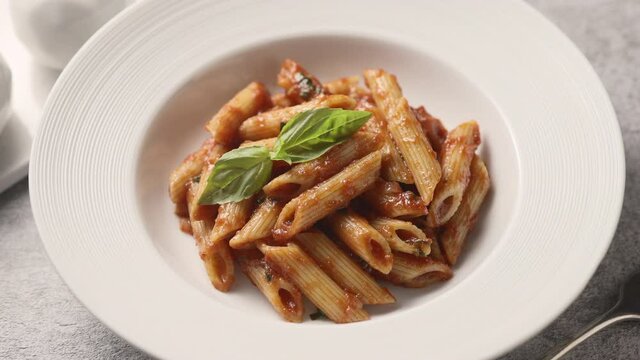 A plate with penne pasta, tomato sauce and basil on a light table, close-up. The camera moves along 