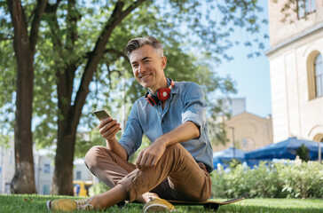 Portrait of handsome smiling man holding mobile phone, looking at camera, sitting on grass. Stylish...