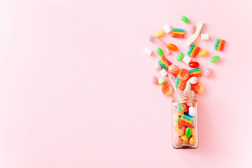 Mixed colorful candies scattered from glass bottle on pink background. Top view