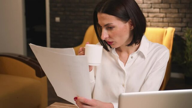 Focused businesswoman drinking coffee, working with papers at home or office. Portrait of woman freelancer reading documents looking on financial statistics diagrams at remote workplace.