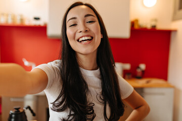 Sincere girl in white T-shirt laughs, looks into camera and takes selfie in kitchen