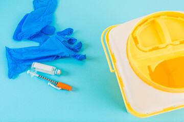 Yellow medical waste container next to blue syringe, vaccine and latex gloves on a blue background