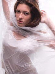 conceptual portrait, one young beautiful woman wraped in plastic sheet. Shot in studio, white background.
