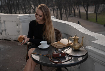 Beautiful smiling girl, with long blond hair, sits at a table with a book and coffee with croissants in her hands