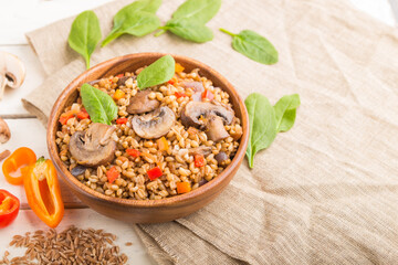 Spelt (dinkel wheat) porridge with vegetables and mushrooms in wooden bowl on a white wooden background and linen textile. Side view, selective focus.